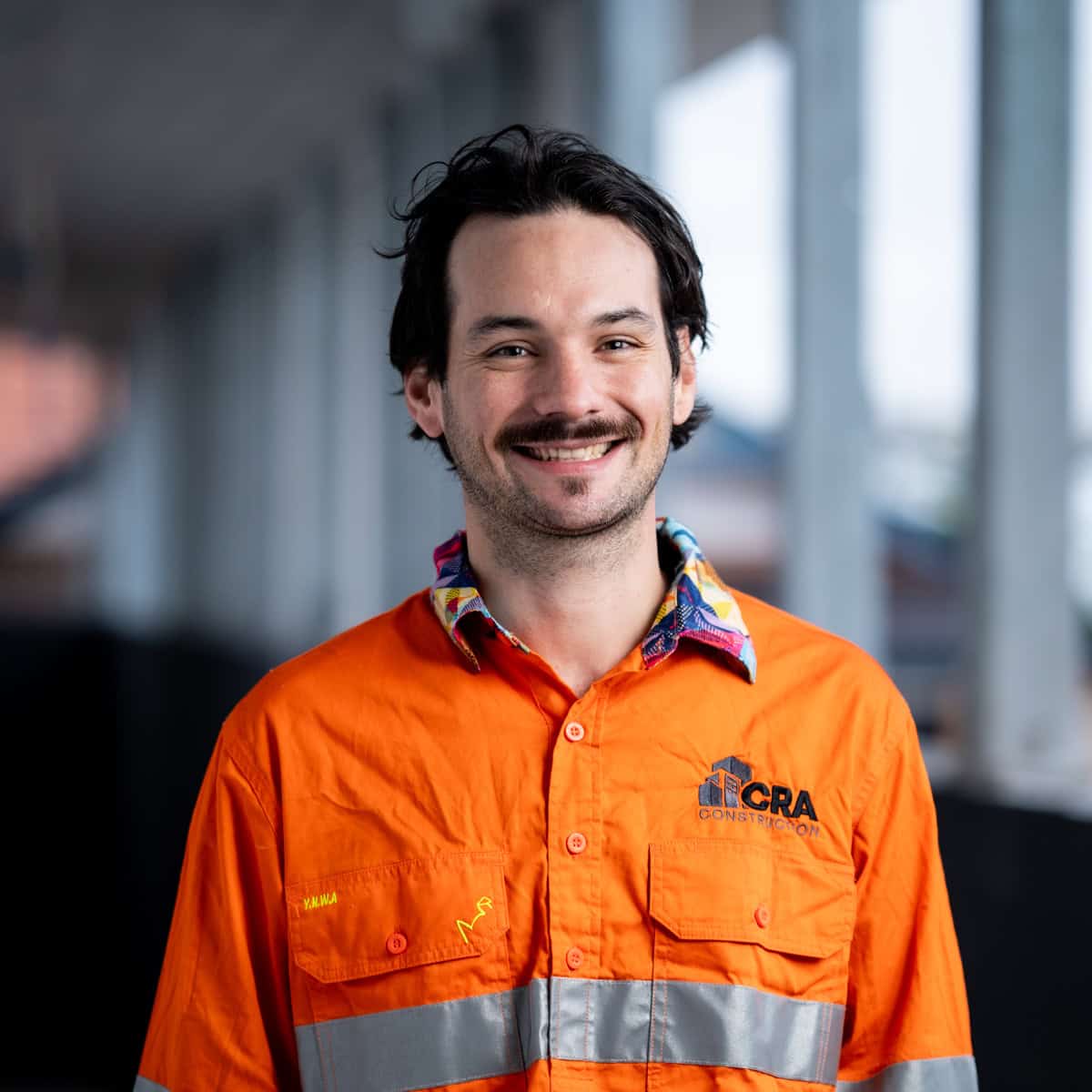 A man with a mustache and black hair, wearing an orange high-visibility construction uniform with CRA Constructions embroidered on it, stands indoors smiling. This proud member of our Plastering Specialists team exudes professionalism and expertise.