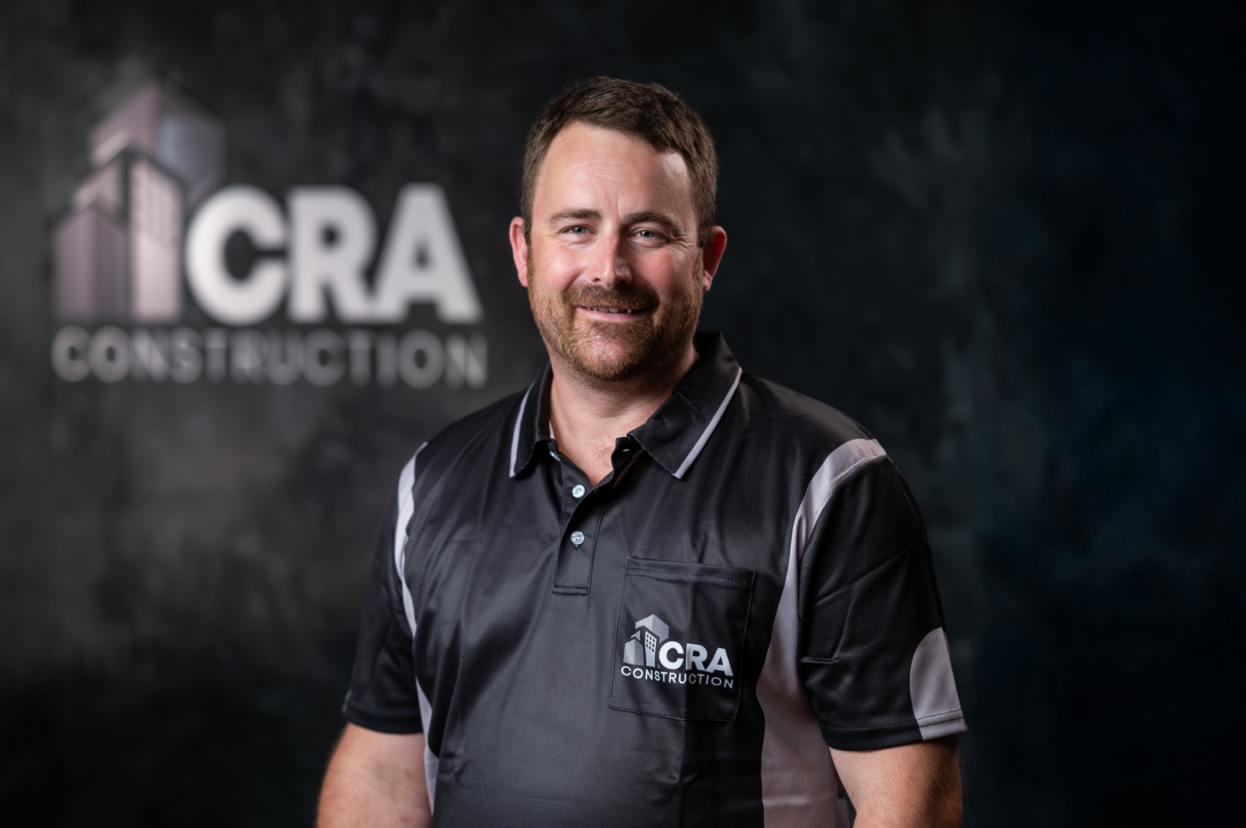 A man in a shirt with "CRA Construction" stands in front of a dark background featuring the company logo. Known for being plastering specialists, CRA Construction prides itself on exceptional craftsmanship and quality.