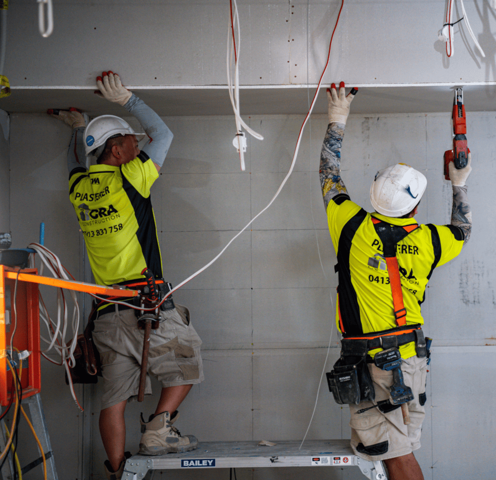 Two Brisbane construction workers in yellow vests and hard hats install drywall indoors, with one using a screw gun and the other holding a tool.