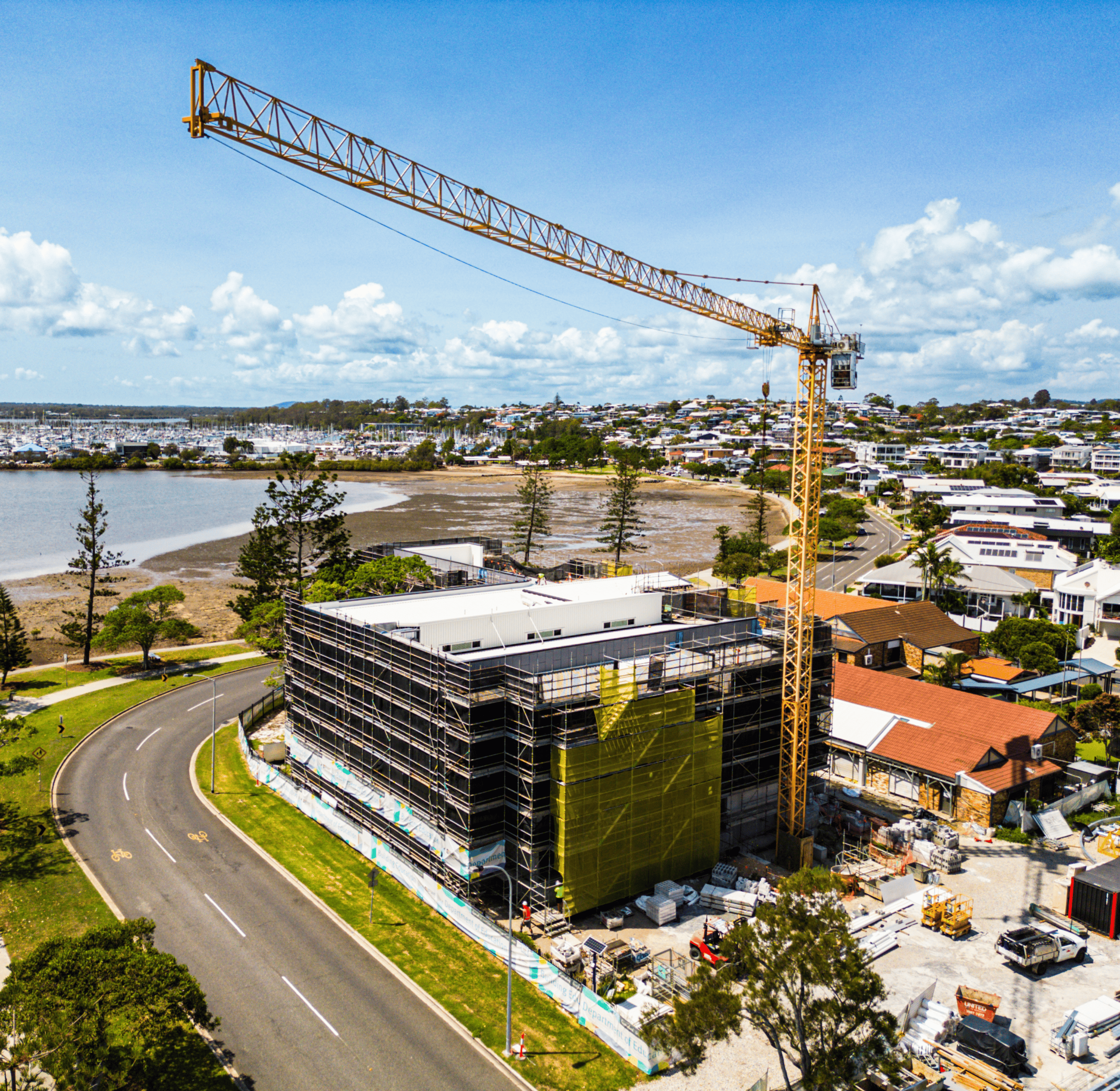 An aerial view shows a yellow construction crane over a multi-story building undergoing construction. Scaffolding surrounds the building, which is partially covered in yellow netting. In the background, a body of water with boats and a residential area are visible under a blue sky with clouds.