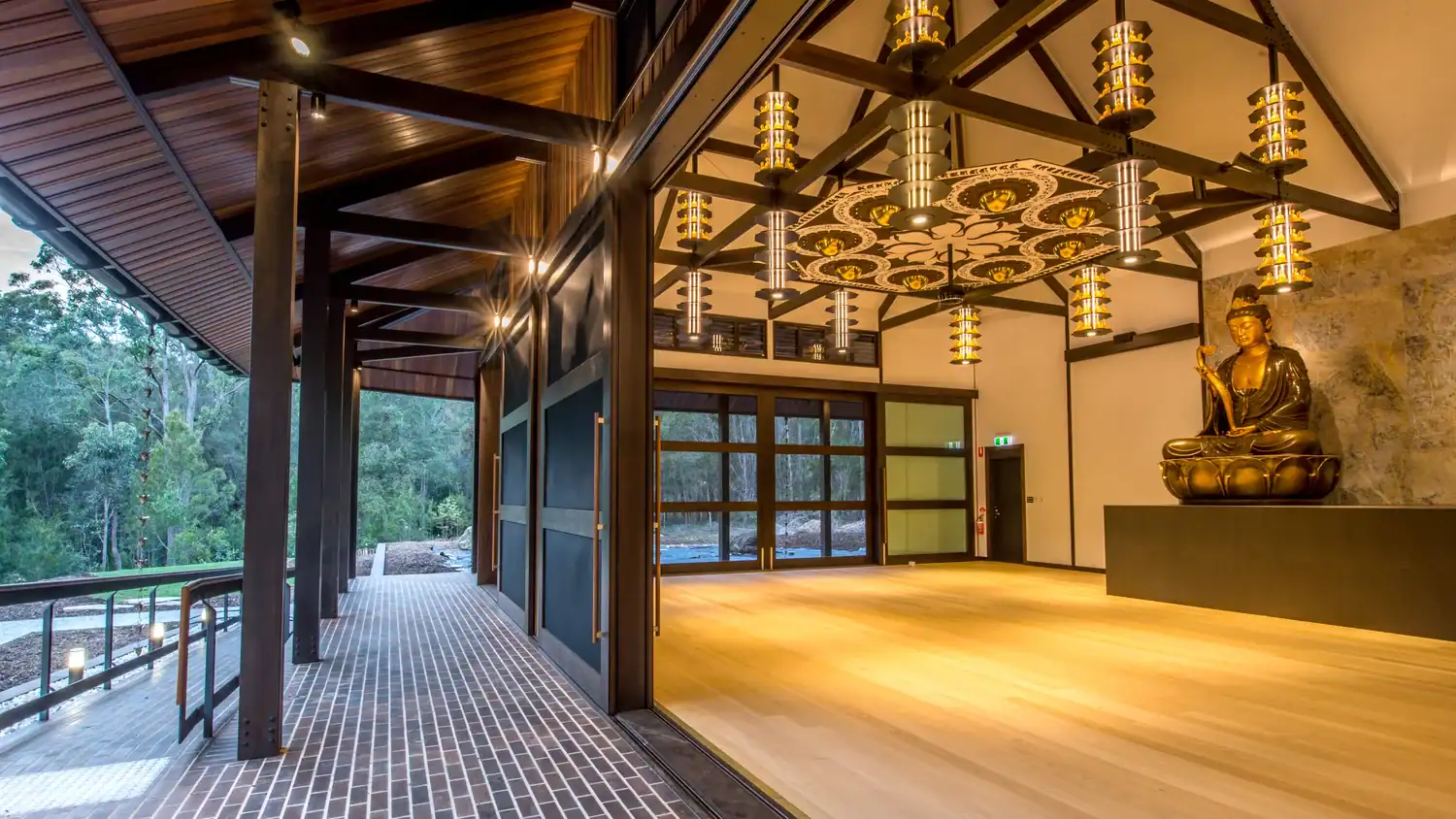 A spacious meditation hall with wooden floors, decorative ceiling lights by commercial plasterers, large windows, and a Buddha statue on a raised platform, surrounded by forest views.