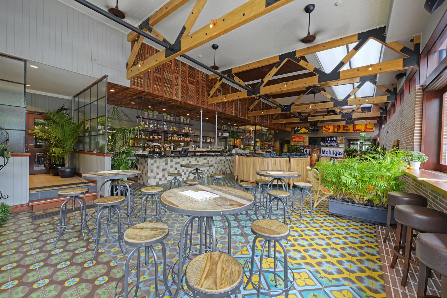 A spacious bar with wooden tables and metal stools, vibrant patterned tile floor, large windows, plants, and exposed beams—perfectly finished by skilled commercial plasterers—with vintage signs on the walls.