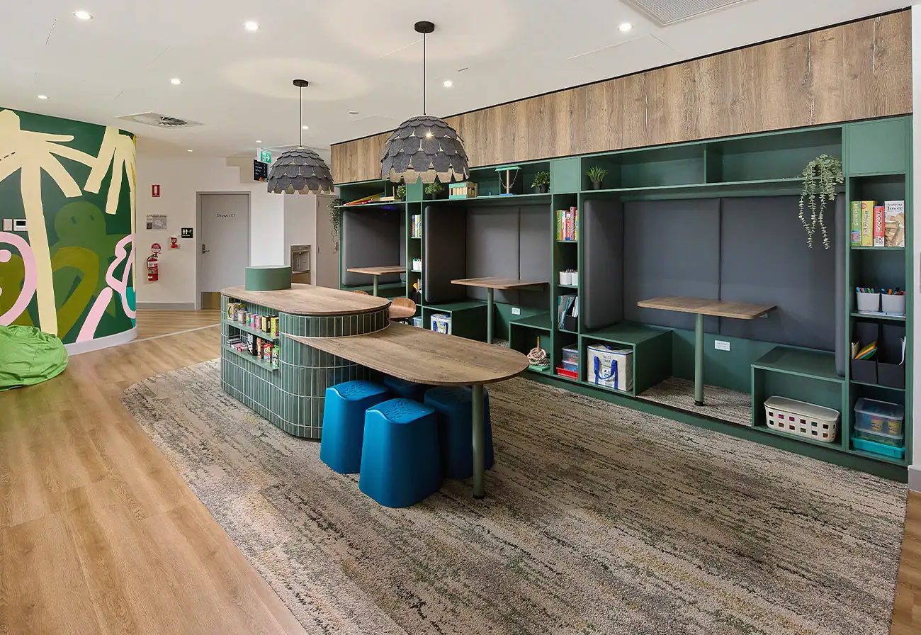 Modern classroom with green built-in shelves, tables, blue stools, educational materials, and a colorful abstract mural on the wall expertly finished by commercial plasterers.