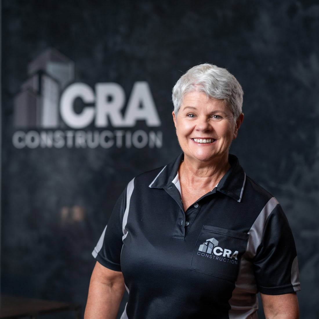 A woman with short gray hair wearing a CRA Construction polo shirt stands smiling in front of a wall with the CRA Construction logo, proudly representing the team of Plastering Specialists.