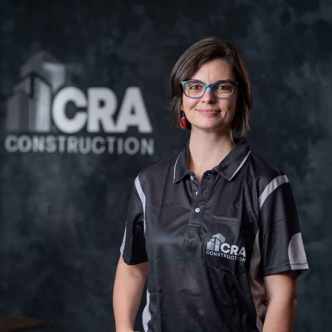 A woman wearing glasses and a CRA Construction shirt stands in front of a wall with the CRA Construction logo, representing the team of Plastering Specialists.