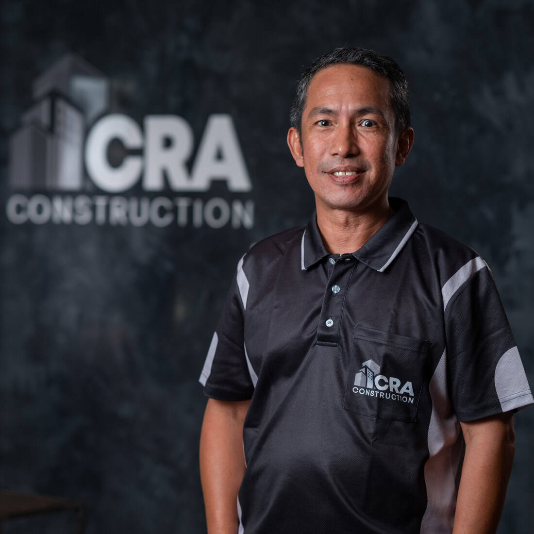 A man wearing a black CRA Construction polo shirt stands in front of a wall with the CRA Construction logo, representing the trusted plastering specialists.