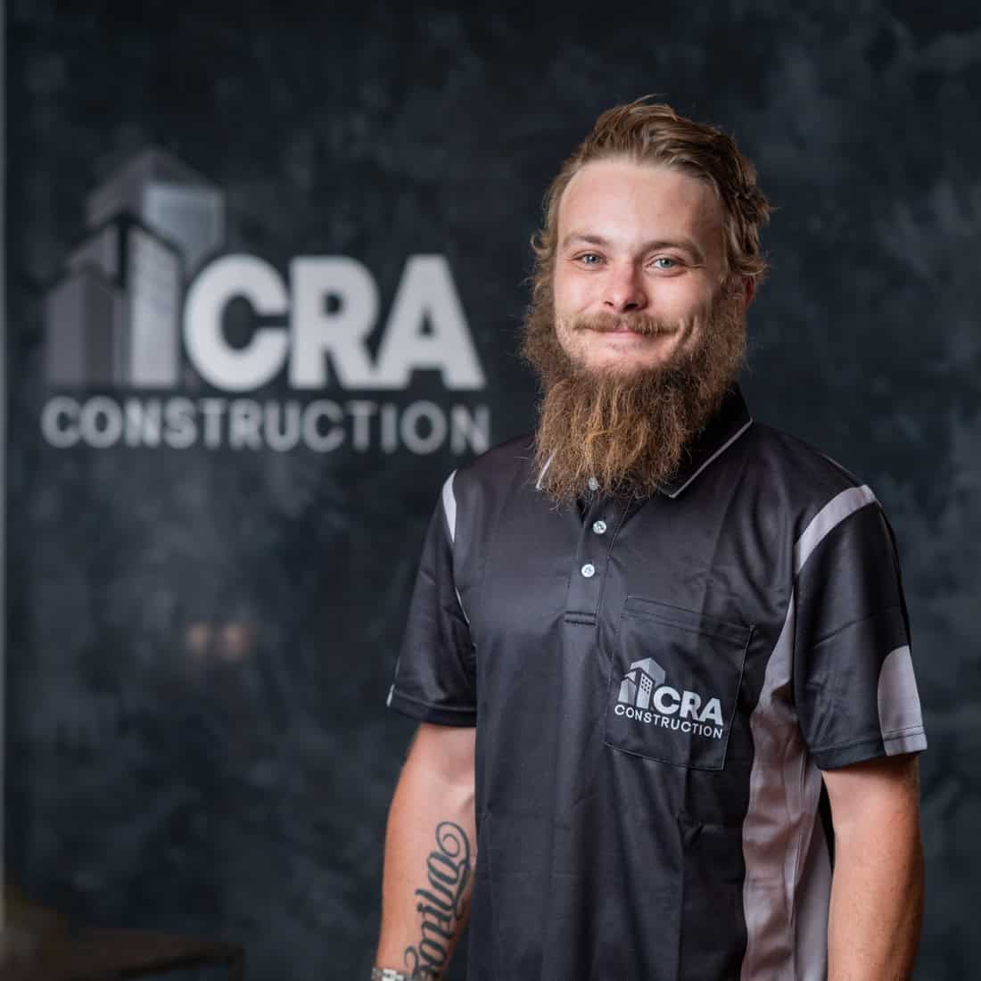 A man with a beard wearing a CRA Construction polo shirt stands in front of a wall with the CRA Construction logo, representing the team of plastering specialists.