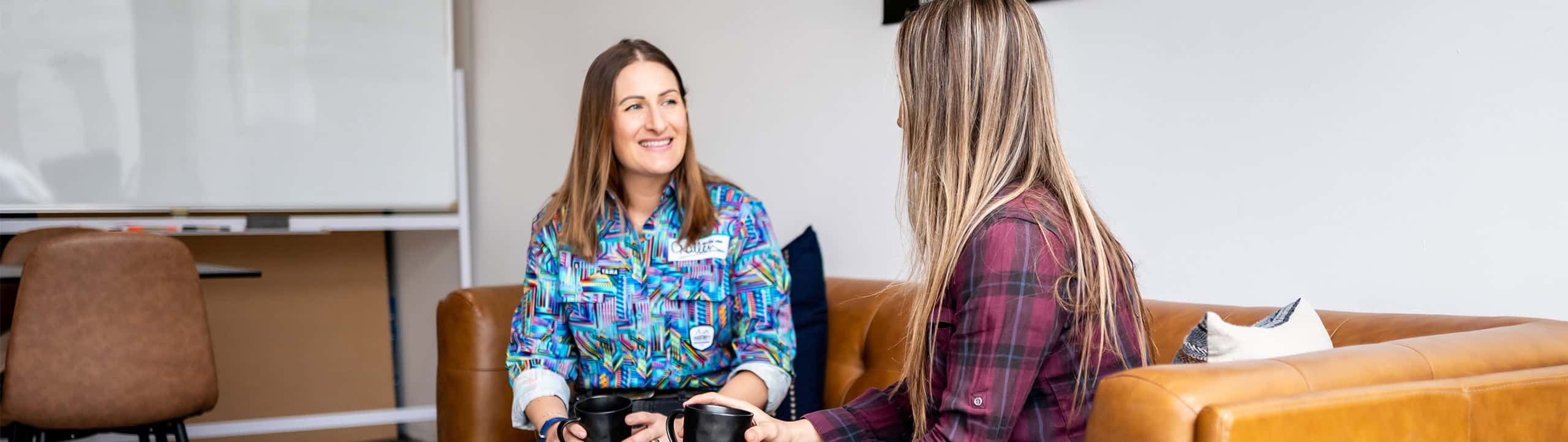 Two women sit on a brown couch, facing each other and holding black mugs, engaged in conversation in a casual indoor setting.